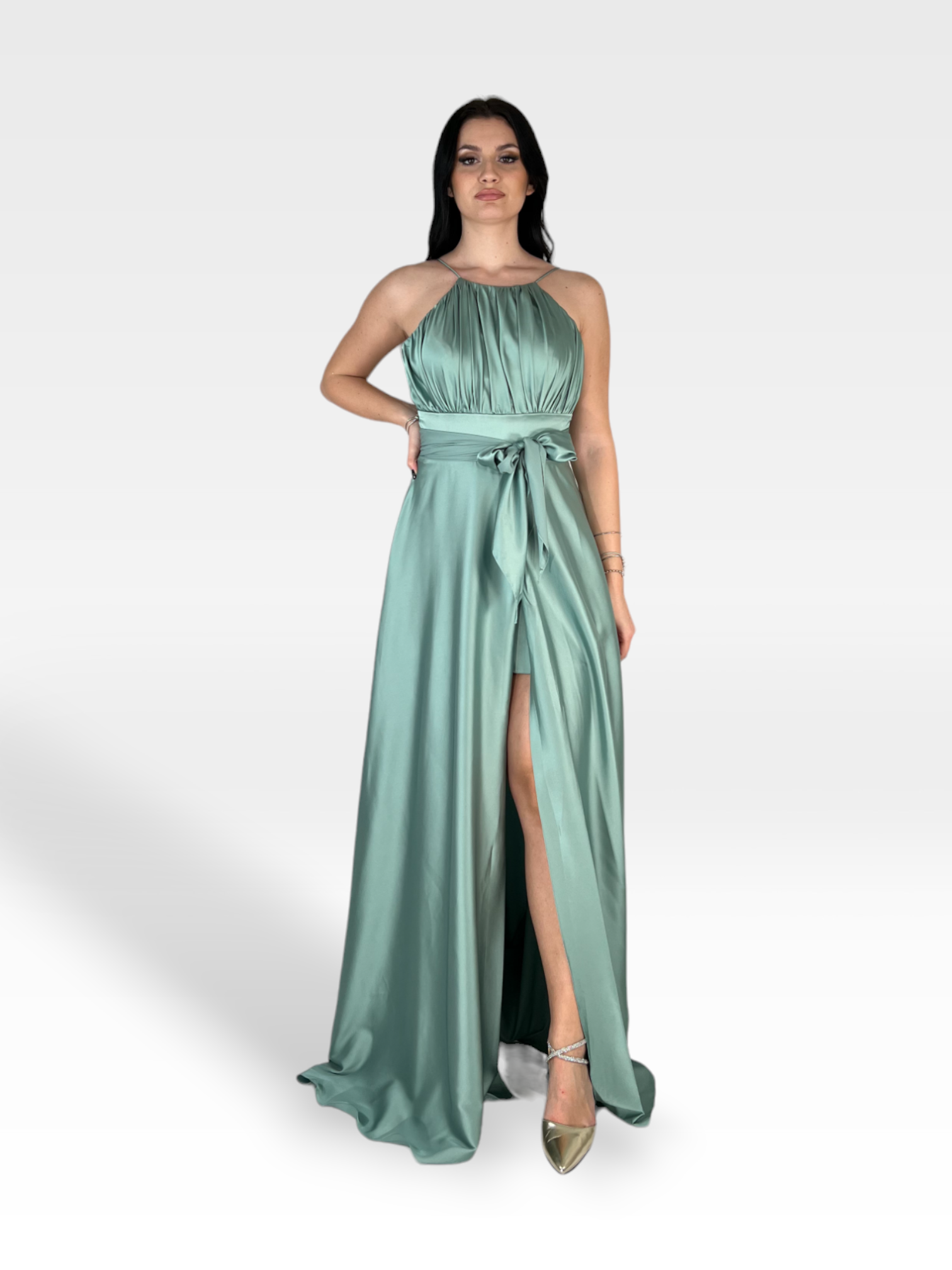 Una donna indossa con sicurezza un abito ELVENE fluido, senza maniche, verde menta, con uno spacco alto e scollo all'americana, legato in vita con una fascia abbinata. Indossa scarpe metalliche a punta e ha una mano appoggiata sul fianco. Il semplice sfondo bianco mette in risalto il suo abbigliamento elegante da 18 anni.