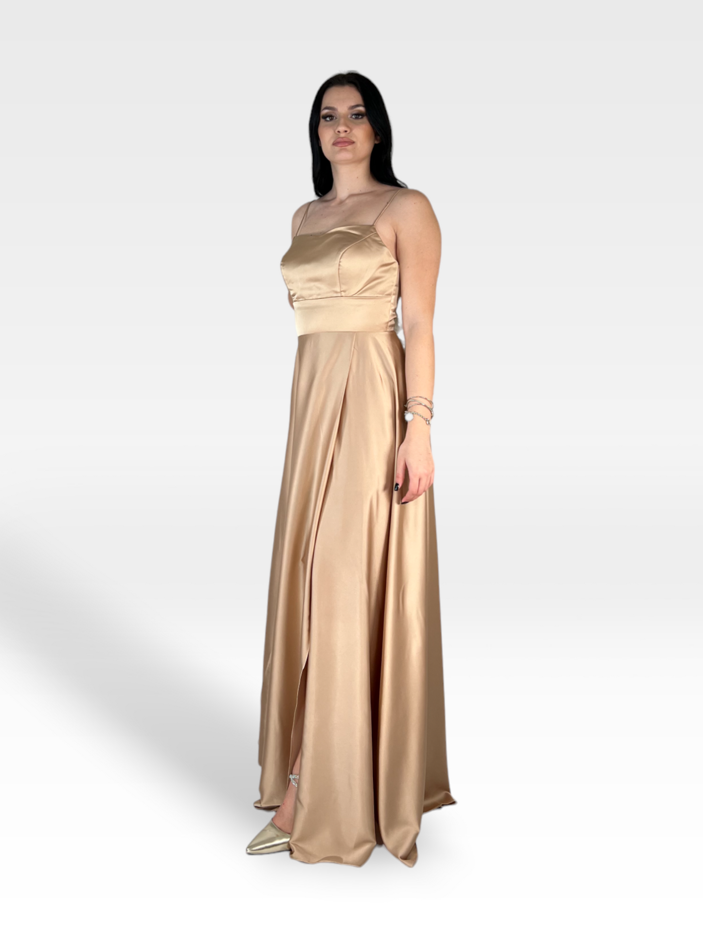 Una donna con lunghi capelli scuri indossa un abito IMMAN in raso color champagne lungo fino al pavimento di 18teen con spalline sottili, che irradia eleganza senza tempo. Lei sta in una posizione neutra su un semplice sfondo bianco, accessoriata con braccialetti e ballerine dorate.