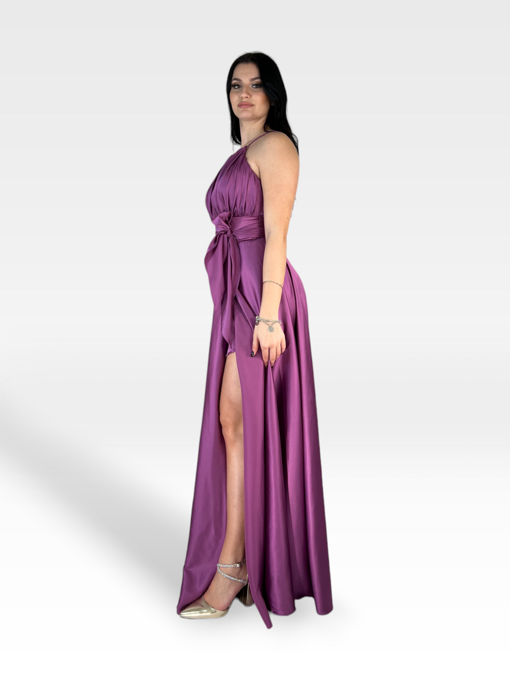 Una donna con lunghi capelli scuri posa con un abito di raso VIOLA senza maniche, lungo fino al pavimento, con spacco laterale e scollo all'americana di 18teen. Indossa tacchi alti argentati e sta in piedi su uno sfondo semplice, grigio chiaro.