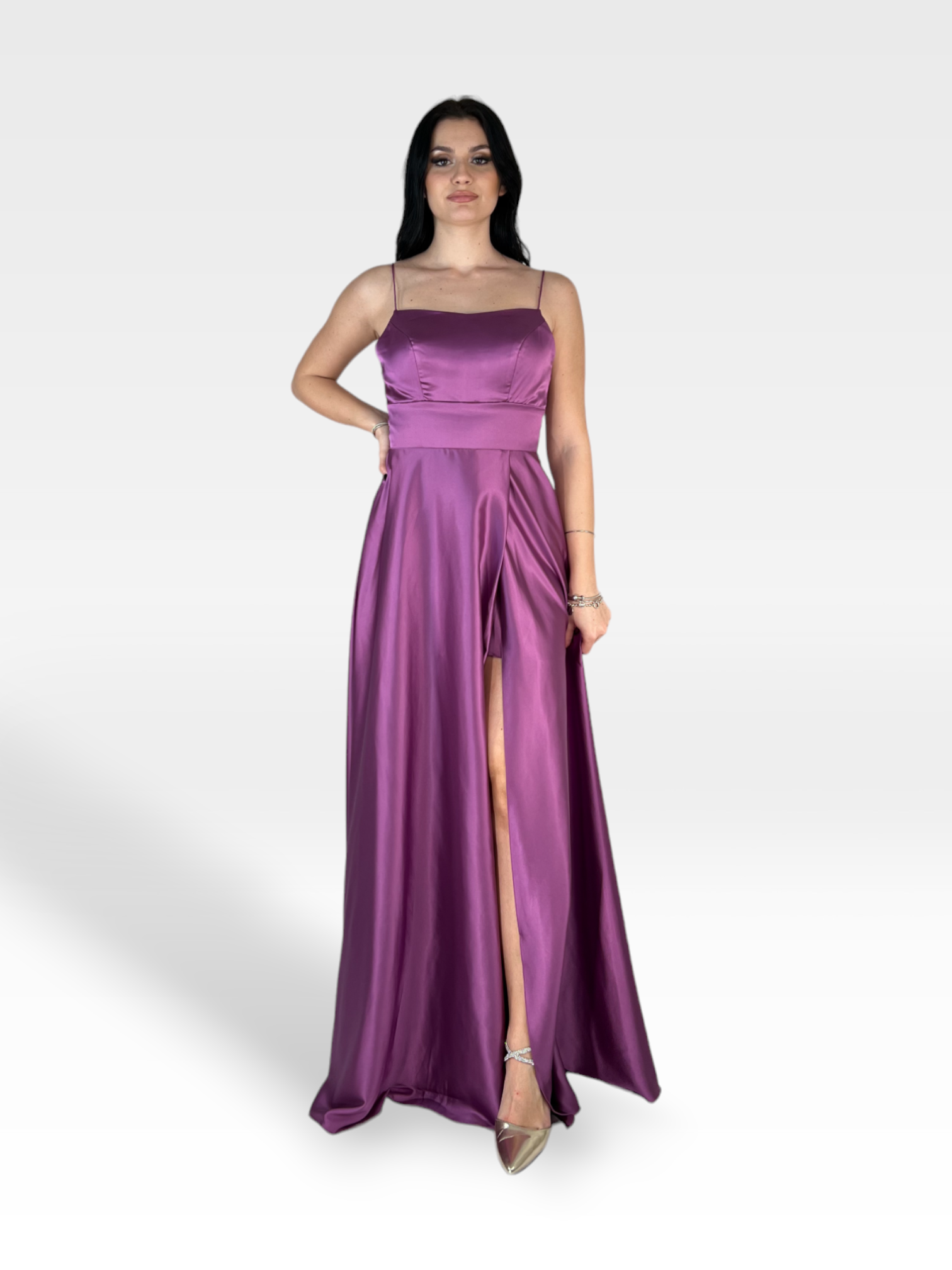 Una donna in piedi su uno sfondo grigio chiaro, indossa un elegante ALYSSUM by 18teen - un abito lungo in raso magenta con spalline sottili e un alto spacco laterale. Ha una mano sul fianco e indossa scarpe metalliche col tacco alto.