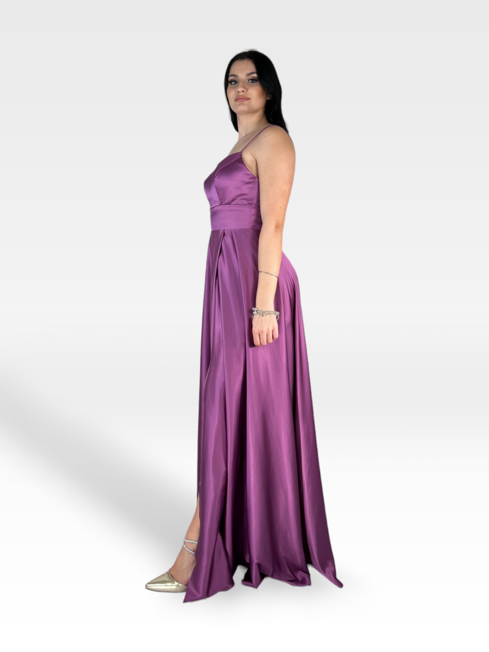 Una donna con lunghi capelli scuri sta in piedi su un semplice sfondo bianco, indossando un abito ALYSSUM in raso viola senza maniche, lungo fino al pavimento, di 18teen con un corpetto aderente e una gonna fluida con uno spacco laterale. Ha un braccio appoggiato al suo fianco e guarda direttamente davanti a sé. Indossa anche i tacchi alti dorati.