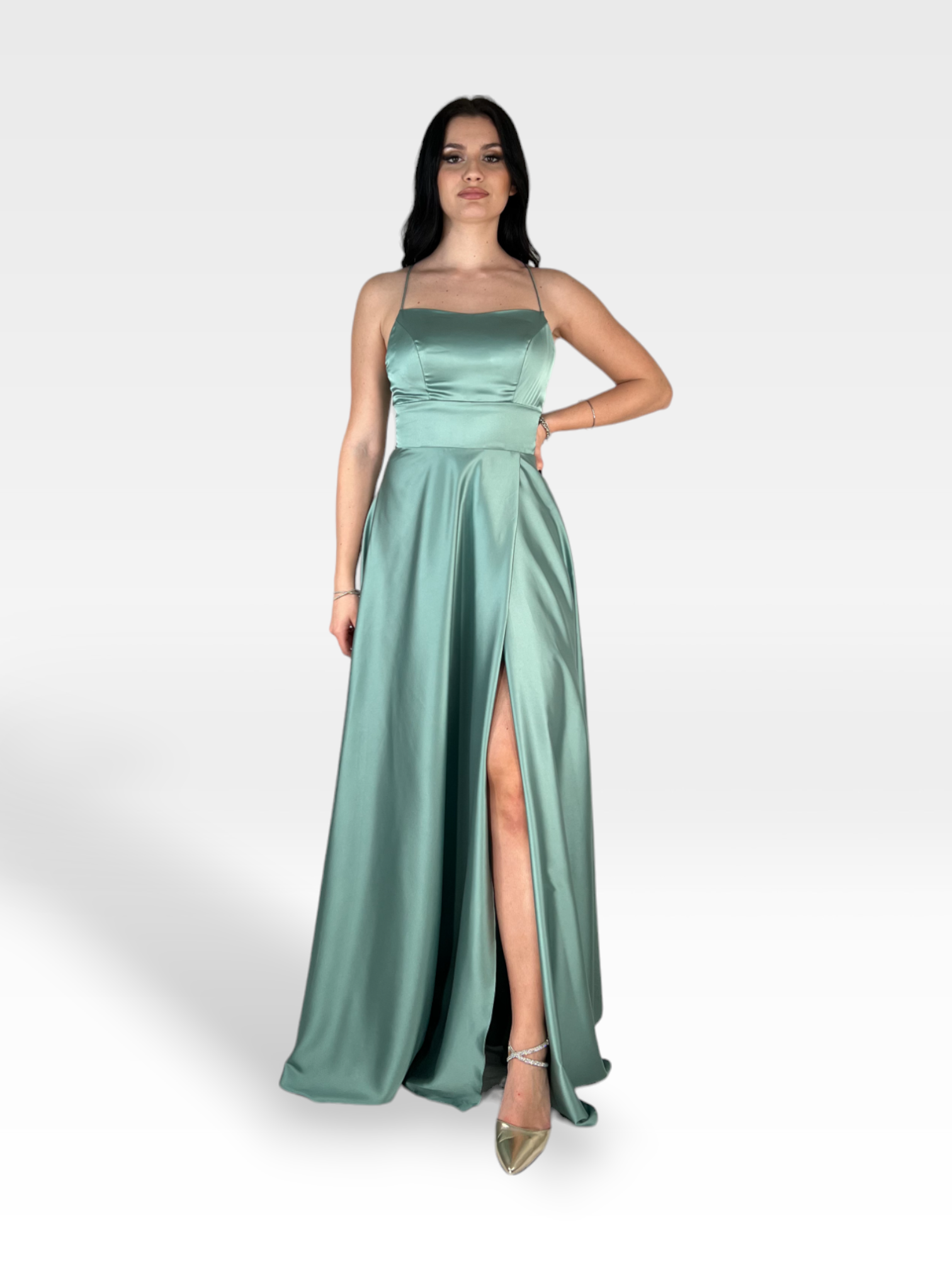 Una donna che indossa una NIENNA 18enne lunga fino al pavimento in raso verde acqua con spalline sottili e uno spacco alto. I suoi lunghi capelli scuri sono raccolti e lei posa con una mano sul fianco. Indossa anche tacchi a spillo argentati. Lo sfondo è un semplice grigio chiaro.