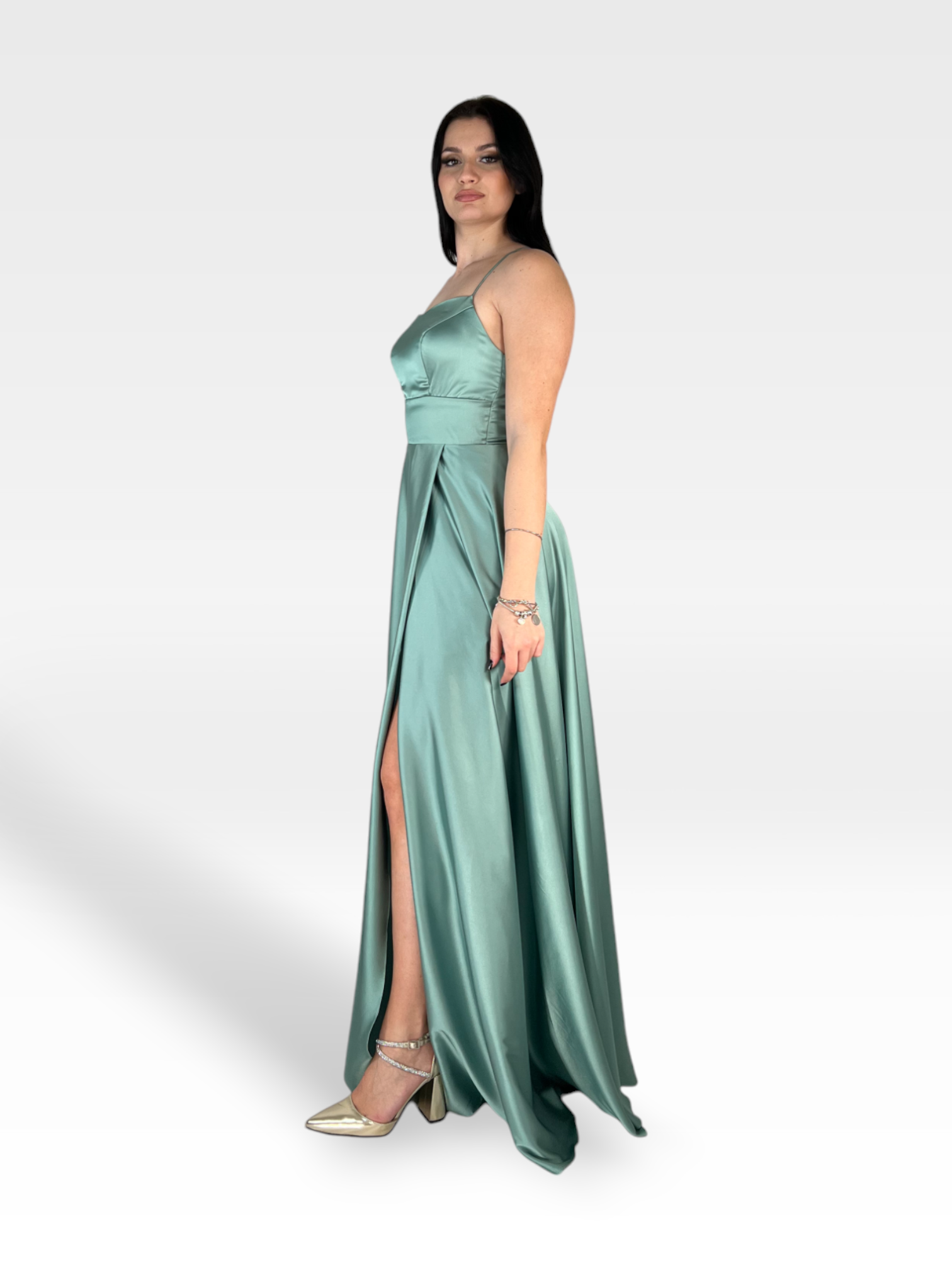 Una donna con un lungo e fluido abito NIENNA di 18teen realizzato in raso verde acqua con spalline sottili posa su uno sfondo semplice grigio chiaro. Indossa tacchi alti dorati e ha uno spacco nel vestito che lascia intravedere una gamba. I suoi lunghi capelli scuri scendono a cascata mentre si accessoria con braccialetti su entrambi i polsi.