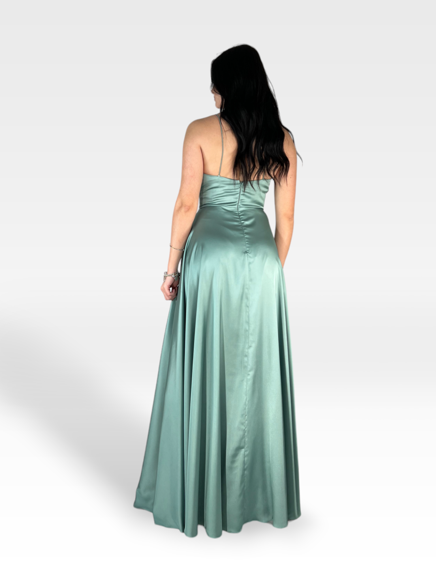 Una donna con lunghi capelli scuri è rivolta lontano dalla telecamera e indossa una NIENNA lunga fino al pavimento e senza spalline in raso verde salvia di 18teen. Lo sfondo è semplice e grigio chiaro, evidenziando il colore salvia e la trama del vestito.