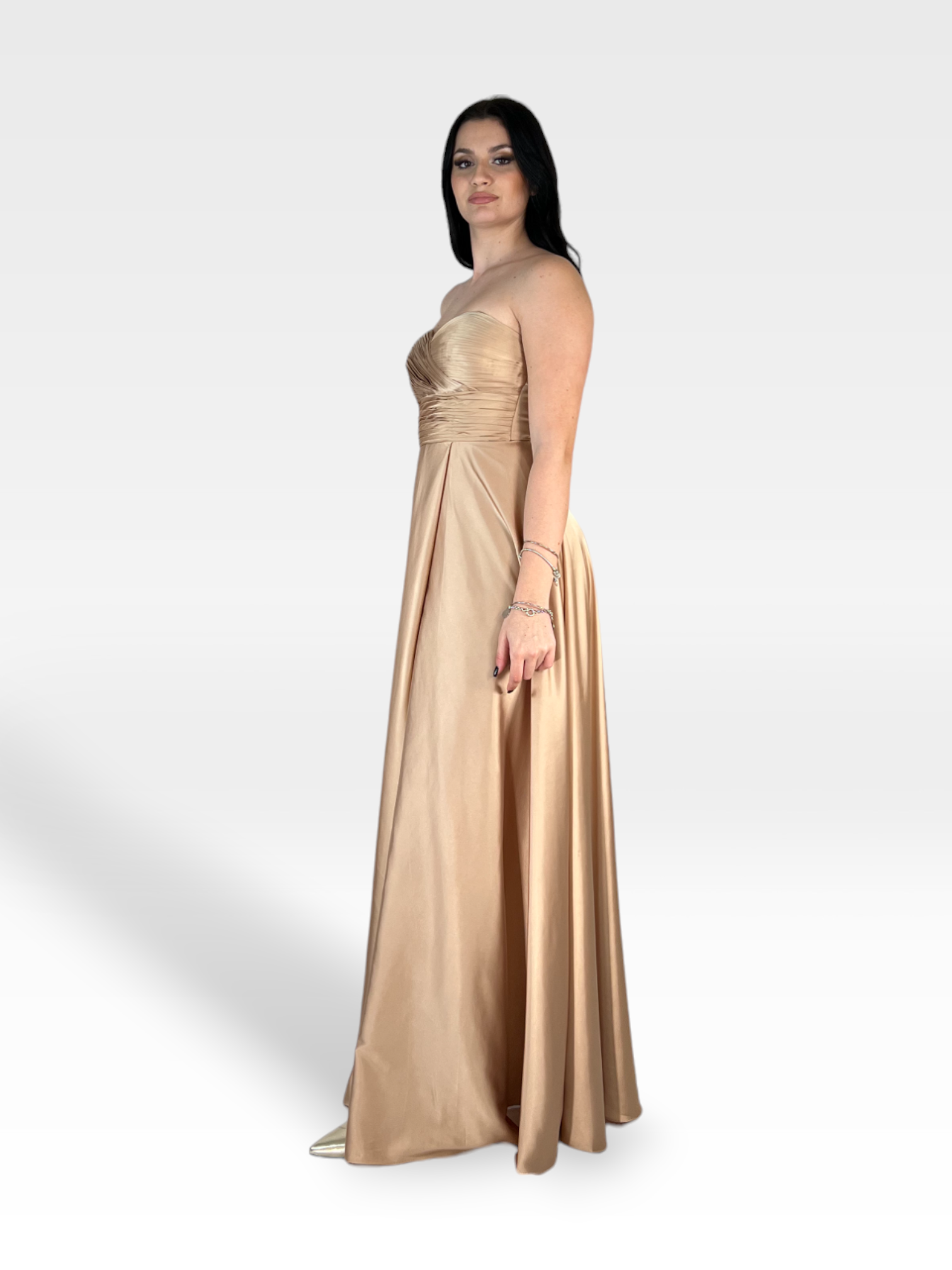 Una donna che indossa un LAKME in raso oro chiaro senza spalline, lungo fino al pavimento, di 18teen posa su un semplice sfondo bianco. Ha lunghi capelli scuri ed è adornata con gioielli minimali, trasudando eleganza senza tempo. Il suo sguardo è diretto verso la telecamera.
